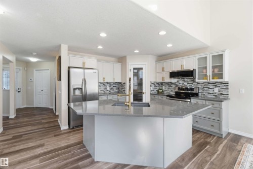 Kitchen featuring a large island with a sink and gold-toned faucet, stainless steel refrigerator and range, tiled backsplash, and white and gray cabinetry with gold-toned hardware - 31 Everitt Drive, St. Albert, AB - Indoor Photo Showing Kitchen With Upgraded Kitchen