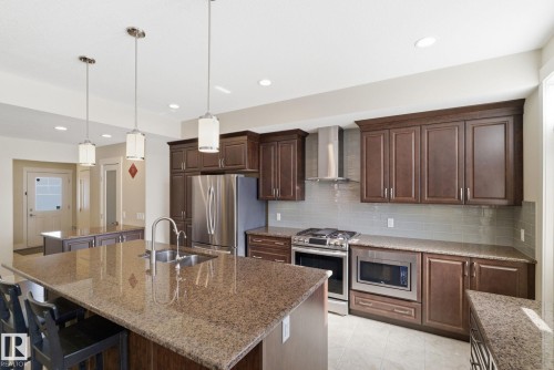 The kitchen features a large island with a double sink and pendant lighting, dark wood cabinetry, granite countertops, and stainless steel appliances - 3235 Abbott Crescent, Edmonton, AB - Indoor Photo Showing Kitchen With Double Sink With Upgraded Kitchen
