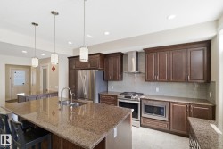 The kitchen features a large island with a double sink and pendant lighting, dark wood cabinetry, granite countertops, and stainless steel appliances - 