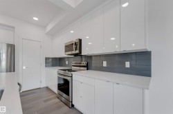 The kitchen features white cabinetry, stainless steel appliances, a dark grey tiled backsplash, and light countertops - 