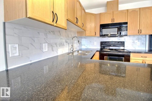 The kitchen features light wood cabinetry, a black oven and microwave, a stainless steel sink with a faucet, a white tile backsplash, and dark speckled countertops - 209 10235 112 Street, Edmonton, AB - Indoor Photo Showing Kitchen With Double Sink