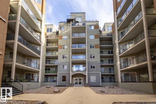 The property features a multi-story building with a neutral-toned exterior, glass-paneled balconies, and a prominent arched entrance - 209 10235 112 Street, Edmonton, AB - Outdoor With Balcony With Facade