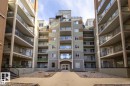 The property features a multi-story building with a neutral-toned exterior, glass-paneled balconies, and a prominent arched entrance - 209 10235 112 Street, Edmonton, AB  - Outdoor With Balcony With Facade 