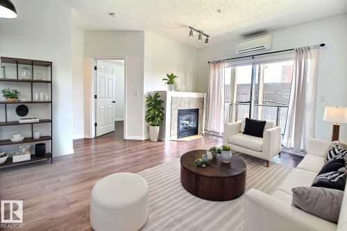 Living area with wood-style flooring, a corner fireplace featuring tile surround, and sliding glass doors with curtains. Virtually Staged - 209 10235 112 Street, Edmonton, AB - Indoor Photo Showing Living Room With Fireplace