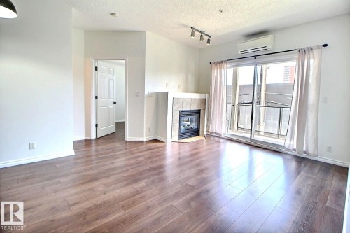Living area featuring hardwood flooring, a corner fireplace with tile surround, and a sliding glass door leading to a balcony - 209 10235 112 Street, Edmonton, AB - Indoor Photo Showing Other Room With Fireplace