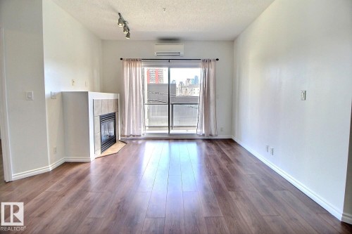 Spacious living area featuring dark wood flooring, a corner fireplace with tile surround, and a sliding glass door with curtains leading to a balcony - 209 10235 112 Street, Edmonton, AB - Indoor With Fireplace