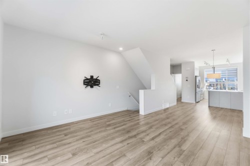Bright living area featuring light wood-style flooring, white walls, and a view into the kitchen area with a large window - 49 100 Jensen Lakes Boulevard, St. Albert, AB - Indoor