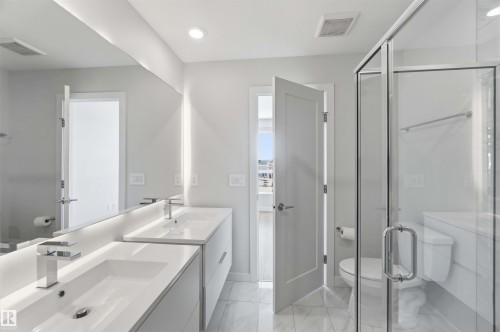 This bathroom features a double vanity with undermount sinks and contemporary faucets, a large wall-mounted mirror with integrated lighting, a glass-enclosed shower, and tiled flooring - 49 100 Jensen Lakes Boulevard, St. Albert, AB - Indoor Photo Showing Bathroom