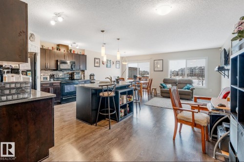 This open-concept living area features rich hardwood flooring, a well-appointed kitchen with dark cabinetry and stainless steel appliances, and an island with seating - 3109 Carpenter Landing, Edmonton, AB - Indoor Photo Showing Kitchen With Upgraded Kitchen