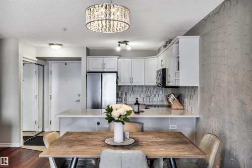 Kitchen and dining area featuring a modern chandelier, white cabinetry, stainless steel appliances, a tiled backsplash, and hardwood flooring - 319 17404 64 Avenue, Edmonton, AB - Indoor Photo Showing Dining Room