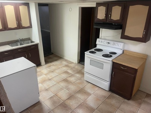 The kitchen features a white electric range, brown cabinetry with light-colored inserts, a double basin sink, and tiled flooring - 12335 132 Avenue, Edmonton, AB - Indoor Photo Showing Kitchen With Double Sink