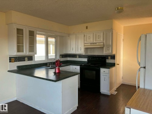 Kitchen featuring white cabinetry, dark countertops, dark wood-finish flooring, a white tile backsplash with dark accent tiles, and a built-in oven and range - 4129 134A Avenue Nw, Edmonton, AB - Indoor Photo Showing Kitchen With Double Sink