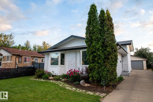 The property features a well-maintained lawn, a paved driveway, and a front garden with mature trees and blooming red flowers - 2626 49 Street, Edmonton, AB - Outdoor