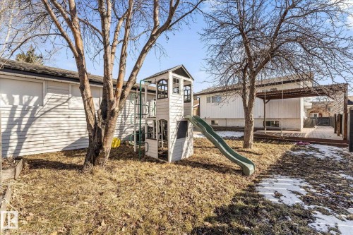 The property includes a spacious yard with mature trees, a wooden deck with an overhead pergola, and a white play structure with a green slide - 2626 49 Street, Edmonton, AB - Outdoor