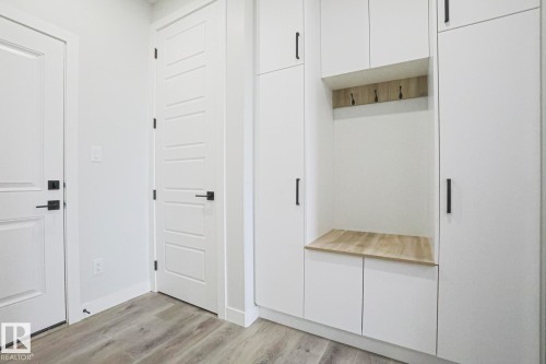 Entryway with light wood tone flooring, white paneled doors with black hardware, and integrated white cabinetry with a wood-look bench and coat hooks - Rural Leduc County, AB - Indoor Photo Showing Other Room