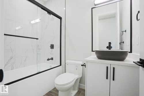 Bathroom featuring a white bathtub with a clear glass sliding shower door and black trim, a toilet, and a vanity with a dark gray vessel sink and black faucet - Rural Leduc County, AB - Indoor Photo Showing Bathroom