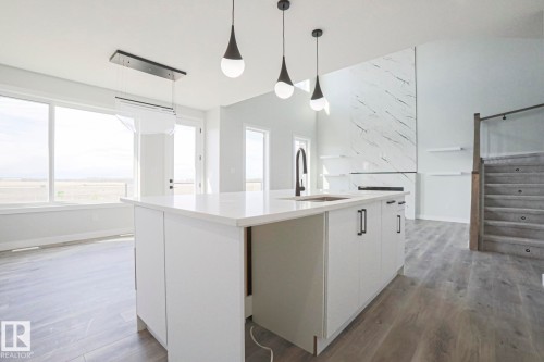 The open-concept living space features a kitchen island with a sink and modern pendant lighting, large windows, and a stone fireplace with shelving - Rural Leduc County, AB - Indoor Photo Showing Kitchen