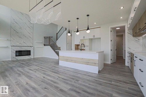 This expansive open-concept living area features light-toned flooring, a grand fireplace with a natural stone surround, and a kitchen island with pendant lighting - Rural Leduc County, AB - Indoor With Fireplace