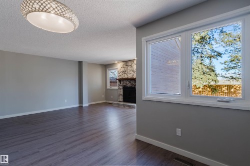 This expansive living area features durable flooring, a decorative overhead light fixture, and large windows that provide natural light and views of trees and a fence - 17308 61 Avenue, Edmonton, AB - Indoor With Fireplace