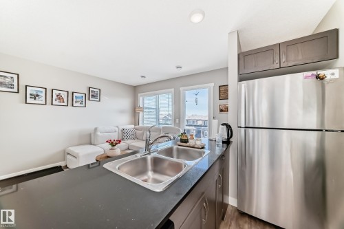 The kitchen features a stainless steel refrigerator, dark wood cabinetry, a double basin sink, and a black countertop - 11 1530 Tamarack Boulevard, Edmonton, AB - Indoor Photo Showing Kitchen With Double Sink
