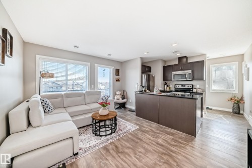 The property features an open-concept living area with large windows, light wood-style flooring, and a kitchen island with a dark countertop, complemented by dark cabinetry and stainless steel appliances - 11 1530 Tamarack Boulevard, Edmonton, AB - Indoor