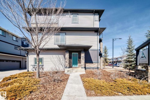 The property features a three-story exterior with siding and stone accents, a prominent front entrance with a teal door, and a paved walkway leading to the entrance - 11 1530 Tamarack Boulevard, Edmonton, AB - Outdoor