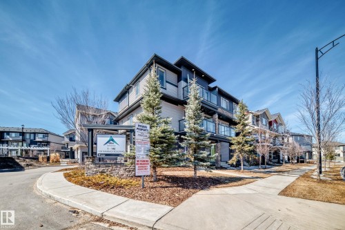 The property features a modern exterior with varied siding, glass-paneled balconies, and a stone-clad sign at the entrance - 11 1530 Tamarack Boulevard, Edmonton, AB - Outdoor With Balcony