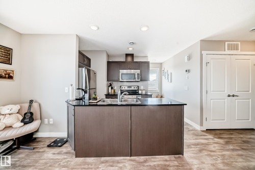 The kitchen features a central island with a sink, dark cabinetry, and stainless steel appliances, including a refrigerator, microwave, and oven - 11 1530 Tamarack Boulevard, Edmonton, AB - Indoor Photo Showing Kitchen With Double Sink