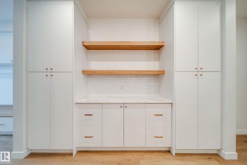Built-in cabinetry featuring white shaker-style doors and drawers with gold-toned hardware, a light-colored countertop, and a white subway tile backsplash, complemented by two floating wooden shelves - 9536 148 Street, Edmonton, AB - Indoor