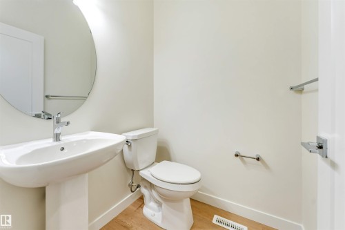 This bathroom features a white pedestal sink with a chrome faucet, a white toilet, a round mirror, and light-toned flooring - 91 Dansereau Way, Beaumont, AB - Indoor Photo Showing Bathroom
