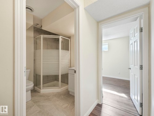 The property features a bathroom with a corner shower enclosure and a white vanity, alongside a separate room with wood-style flooring and a window - 675 Mcallister Loop, Edmonton, AB - Indoor Photo Showing Bathroom