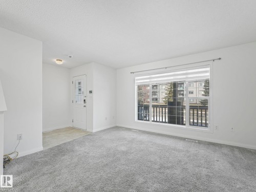 This property features a bright living area with light grey carpeting and a large window, offering views of surrounding buildings and trees - 675 Mcallister Loop, Edmonton, AB - Indoor