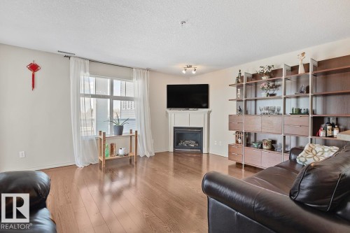 Living area featuring hardwood flooring, a gas fireplace with a mantel, and a large window with white curtains - 225 16035 132 Street, Edmonton, AB - Indoor Photo Showing Living Room With Fireplace