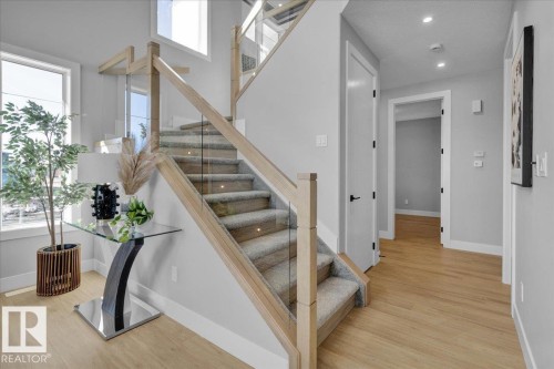 The property features a staircase with light wood railings and glass panels, carpeted stairs with integrated lighting, and light wood flooring in the hallway - 207 Starling, Fort Saskatchewan, AB - Indoor Photo Showing Other Room