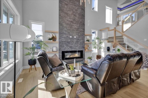 The living area features light-toned flooring, a stone accent wall with a fireplace, and a staircase with glass panels and a wooden handrail - 207 Starling, Fort Saskatchewan, AB - Indoor With Fireplace