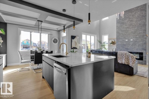 The open concept living area features light wood flooring, a kitchen island with a white countertop and stainless steel dishwasher, and a stone accent wall with a fireplace - 207 Starling, Fort Saskatchewan, AB - Indoor Photo Showing Kitchen With Upgraded Kitchen