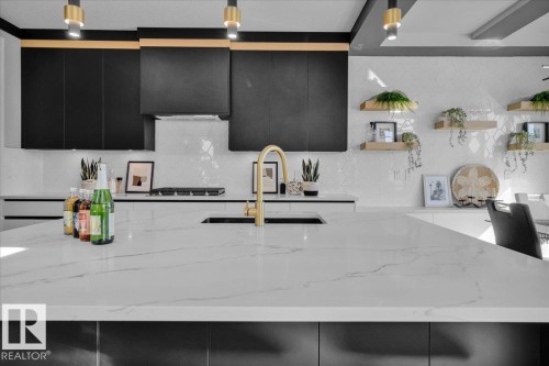 Contemporary kitchen featuring a large island with a sink and a gold-toned faucet, black cabinetry, white countertops, and a white tiled backsplash - 207 Starling, Fort Saskatchewan, AB - Indoor Photo Showing Kitchen With Upgraded Kitchen