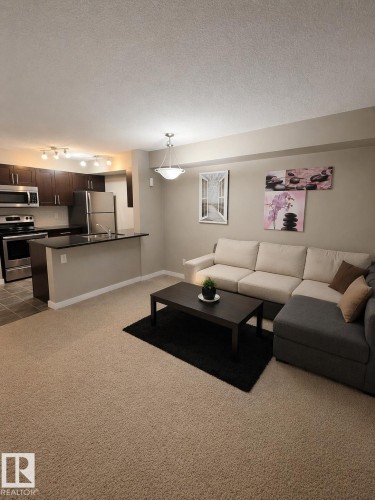 Open-concept living area featuring light-colored carpeting, a kitchen with dark cabinetry, stainless steel appliances, and a breakfast bar - 209 107, Watt Commom, Edmonton, AB 