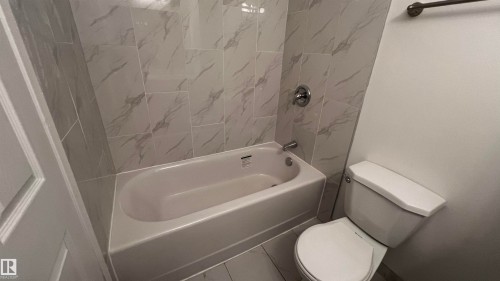 The bathroom features a bathtub with a shower, a toilet, and light-colored wall tiles with a subtle marble pattern - 3323 107 Avenue Nw, Edmonton, AB - Indoor Photo Showing Bathroom