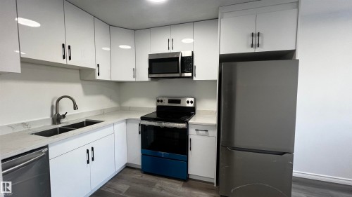 The kitchen features white cabinetry with black hardware, a stainless steel sink with a gooseneck faucet, light-colored countertops, and stainless steel appliances including a refrigerator, microwave, and range - 3323 107 Avenue Nw, Edmonton, AB - Indoor Photo Showing Kitchen With Stainless Steel Kitchen With Double Sink