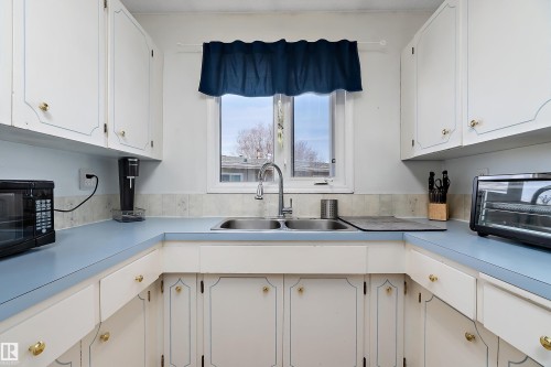 The kitchen features white cabinetry with gold hardware, light blue countertops, and a double basin stainless steel sink beneath a window - 28 Camelot Avenue, Leduc, AB - Indoor Photo Showing Kitchen With Double Sink