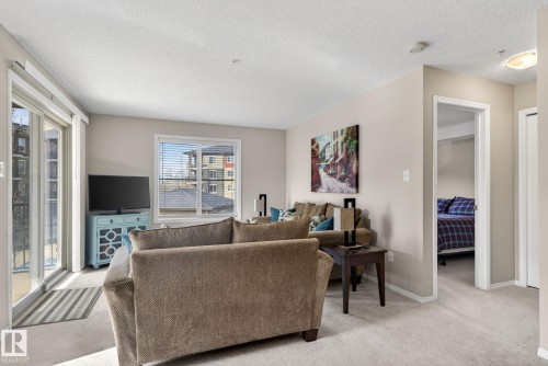The living area features light-colored walls, carpeted flooring, and a window with blinds - 215 1080 Mcconachie Boulevard, Edmonton, AB - Indoor Photo Showing Living Room