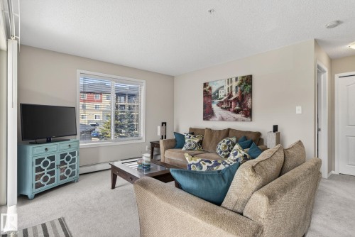 Living area featuring light-colored walls and carpeting, a window with blinds, and a white interior door - 215 1080 Mcconachie Boulevard, Edmonton, AB - Indoor Photo Showing Living Room