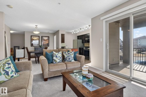 The property features a bright living area with light-colored carpeting, a sliding glass door leading to a balcony, and an open concept layout that includes a dining area and kitchen - 215 1080 Mcconachie Boulevard, Edmonton, AB - Indoor Photo Showing Living Room