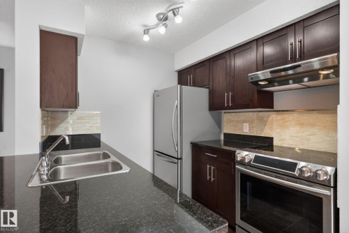 The kitchen features dark wood cabinetry, stainless steel appliances, a double basin sink, and granite countertops - 215 1080 Mcconachie Boulevard, Edmonton, AB - Indoor Photo Showing Kitchen With Double Sink