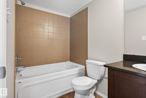 Bathroom featuring a built-in bathtub with shower, a vanity with a dark countertop and white sink, and a toilet - 215 1080 Mcconachie Boulevard, Edmonton, AB - Indoor Photo Showing Bathroom
