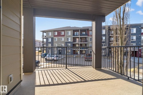 The property features a private balcony with a concrete floor and a black metal railing, providing views of nearby buildings and a paved parking area - 215 1080 Mcconachie Boulevard, Edmonton, AB - Outdoor With Balcony With Exterior