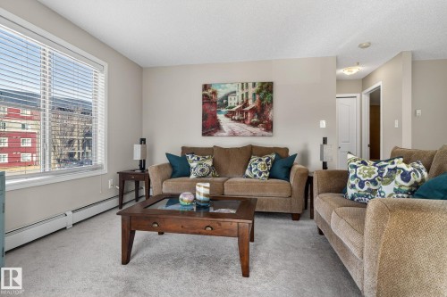 The living area features a large window with blinds, carpeted flooring, and neutral wall tones - 215 1080 Mcconachie Boulevard, Edmonton, AB - Indoor Photo Showing Living Room