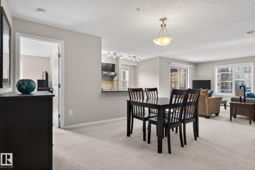 Open concept living area featuring a dining space with a dark wood table and chairs, a kitchen with dark cabinetry and stainless steel appliances, and a living room with a sliding glass door and windows providing natural light - 215 1080 Mcconachie Boulevard, Edmonton, AB - Indoor Photo Showing Dining Room