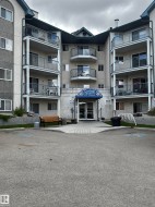 Multi-story residential building featuring multiple balconies, a central entrance with a blue awning, and an asphalt parking area - 